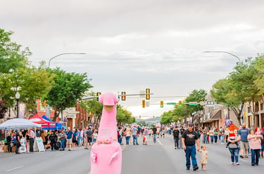 A large pink dinosaur mascot walks down a crowded city street during a summer community festival.