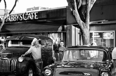 Black and white photo of a cowboy with a vintage Chevrolet car and truck outside Abby's Cafe.
