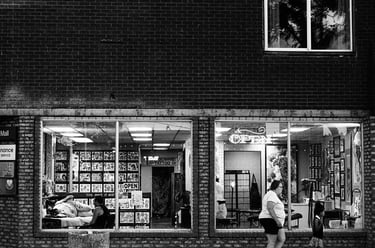 Black and white street photography of people walking past a brick tattoo parlor storefront window.