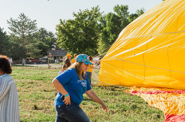 Volunteers prepare a large yellow hot air balloon for flight in a grassy field on a sunny day.