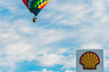 A colorful checkered hot air balloon floats in a blue sky above a Shell gas station price sign.