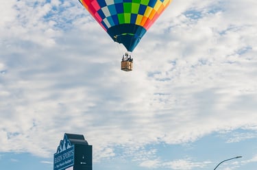 A colorful rainbow checkered hot air balloon flying over a city street with a commercial sign.