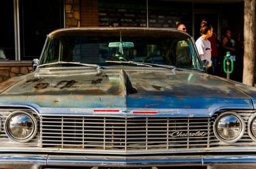 Front view of a classic blue Chevrolet Impala with a rusted hood parked in front of a local salon.