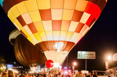 Crowd gathers for a colorful hot air balloon glow event at night with illuminated checkered balloons.