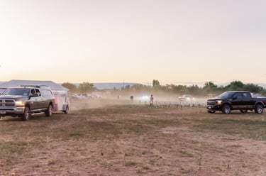 Trucks and trailers parked in a dusty field during an outdoor sunset event with people gathered.