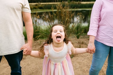 Family photo of a little girl holding her parents hands, she sticks her tongue out at the camera