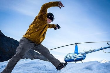 A photographer in hiking gear and crampons leaps across an icy glacier near a blue rescue helicopter.