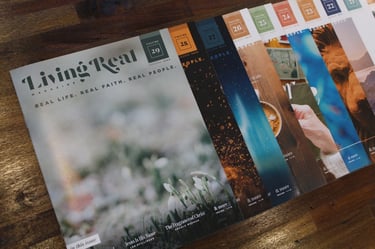 A collection of Living Real Magazine issues fanned out on a wooden table, showcasing faith-based lifestyle content.
