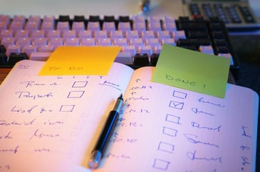 Handwritten to-do list in a notebook with a mechanical keyboard and colorful sticky notes.