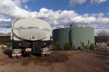 A potable water truck delivery service filling a large green storage tank on a rural landscape.