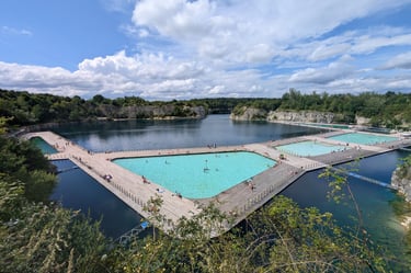 Outdoor swimming pools with turquoise water built on a lake, surrounded by trees and rocky cliffs.