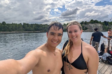 Smiling couple in swimwear taking a selfie by a lake with trees and cloudy sky in the background.