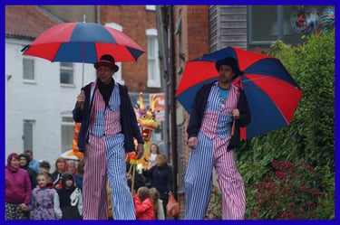 Performers on stilts in striped costumes and red umbrellas walk through a street festival crowd.