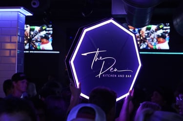 A glowing neon hexagon sign for The Den Kitchen and Bar held up in a crowded nightclub venue.