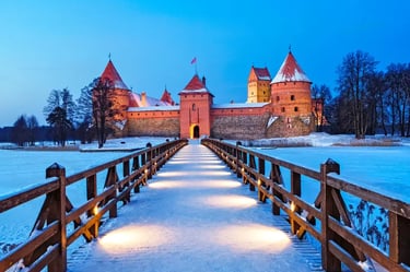 Illuminated Trakai Island Castle in Lithuania at twilight during winter with a snow-covered bridge and frozen lake.