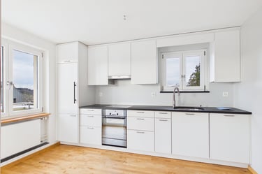 Moderne weiße Küche mit Edelstahlgeräten, schwarzen Arbeitsplatten und hellem Holzboden.