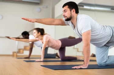 A group of diverse people practicing bird-dog yoga poses on mats in a bright fitness studio.