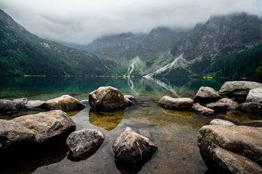 A calm, reflective mountain lake surrounded by rocks and partially shrouded in mist, with trees.