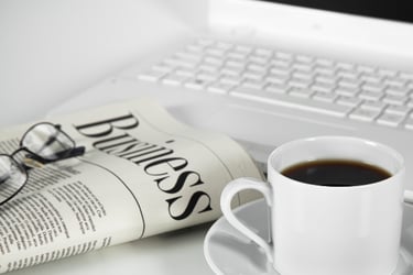 Pic of a folded newspaper with glasses on top, a mug of coffee, and a laptop