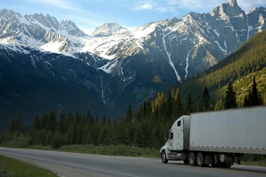 A semi-truck driving on a scenic highway through snow-capped mountains and evergreen forests.
