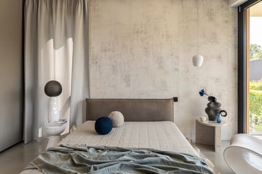 Minimalistyczna jasna sypialnia, ściana o strukturze betonu, beżowe łóżko i designerska lampa.