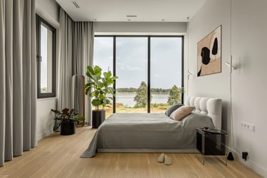 Jasna sypialnia z widokiem, szare tapicerowane łóżko, szklany stolik nocny i obraz abstrakcyjny.