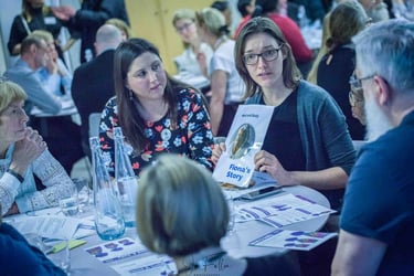 A woman in glasses holding a Fiona's Story pamphlet during a mental health workshop table discussion.