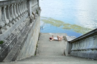 Film still from "Abandonment" by Tina Bikic of a performer lying on wet stone steps beside a lake with green algae.