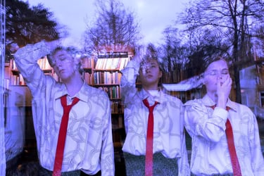 Film still from "Because We Cannot Do Otherwise" by Tina Bikic of three performers in red ties reflected in a large window.