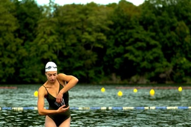 Film still from "Belonging" by Tina Bikic showing a woman standing by the water with trees in the background.