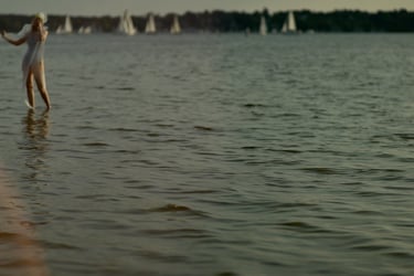 Film still from "Desire for Belonging" by Tina Bikic of a woman dancing through water with sailboats in the distance.