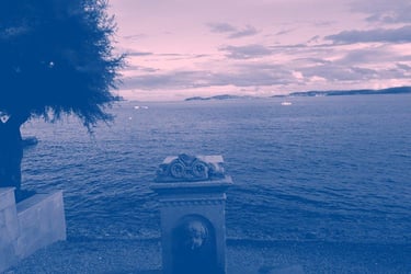 Film still from "Unfinished" by Tina Bikic of a stone fountain by the sea with mountains and boats in the distance.