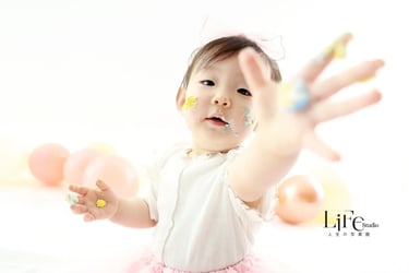 白い背景で、スマッシュケーキのクリームが顔や手についた1歳の女の子が笑顔で写るバースデーフォトの撮影風景