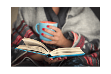 A woman wrapped in a blanket reads a book and holds a mug