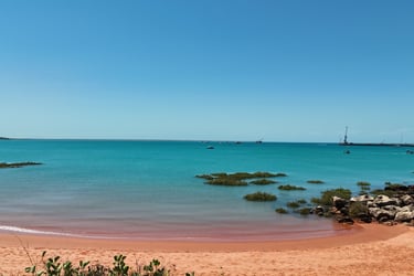 Turquoise ocean water meets red sand at a tropical beach with rocky outcrops and a distant pier.