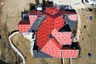 Aerial drone view of professional roofers installing new asphalt shingles over red underlayment on a residential home.
