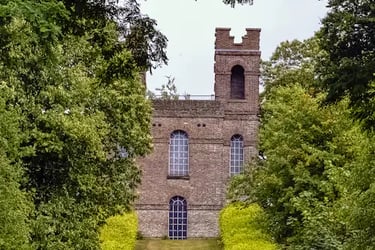 A historic brick folly castle with arched windows framed by lush green trees and a grassy pathway.