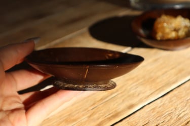 A person holding a small, polished coconut shell bowl on a rustic wooden table with natural sunlight.