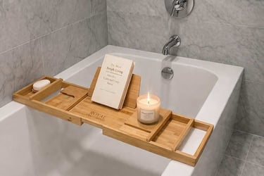 A bamboo bathtub tray caddy holds a book and a candle across a modern white soaking tub.