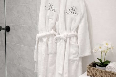 Two white monogrammed robes hang on a bathroom door next to a small orchid and a spa basket