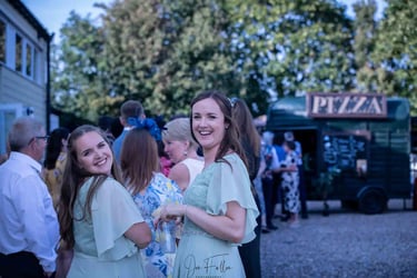 Smiling bridesmaids at an outdoor wedding reception with a horsebox pizza catering van.