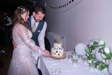 A bride and groom smiling while cutting their two-tier white wedding cake decorated with pressed flowers.