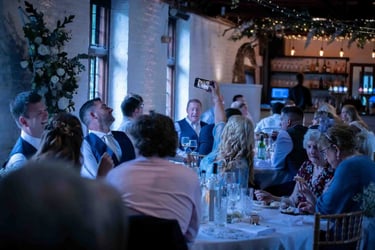 Wedding guests smiling and taking photos with smartphones at a rustic indoor reception dinner.
