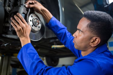 A professional auto mechanic in blue coveralls inspecting a car's brake disc and caliper in a repair shop.