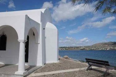 White Greek Orthodox church by the sea with a wooden bench overlooking the Aegean coast.