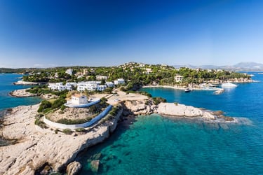Aerial view of a coastal Greek village with white houses, turquoise sea, and rocky cliffs under a clear blue sky.