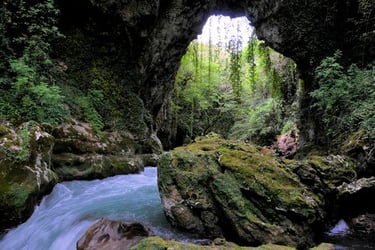 A turquoise river flows through a lush, mossy limestone cave opening in a forest landscape.