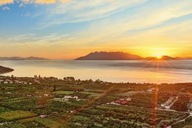 Golden sunset over a coastal village and lush green groves with mountains and calm sea in the background.