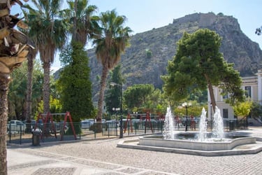 A sunny public square with palm trees and a tiered water fountain below a hilltop fortress.