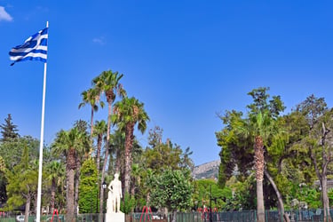 A Greek flag waving in a sunny park with palm trees and a white marble statue in Nafplio.